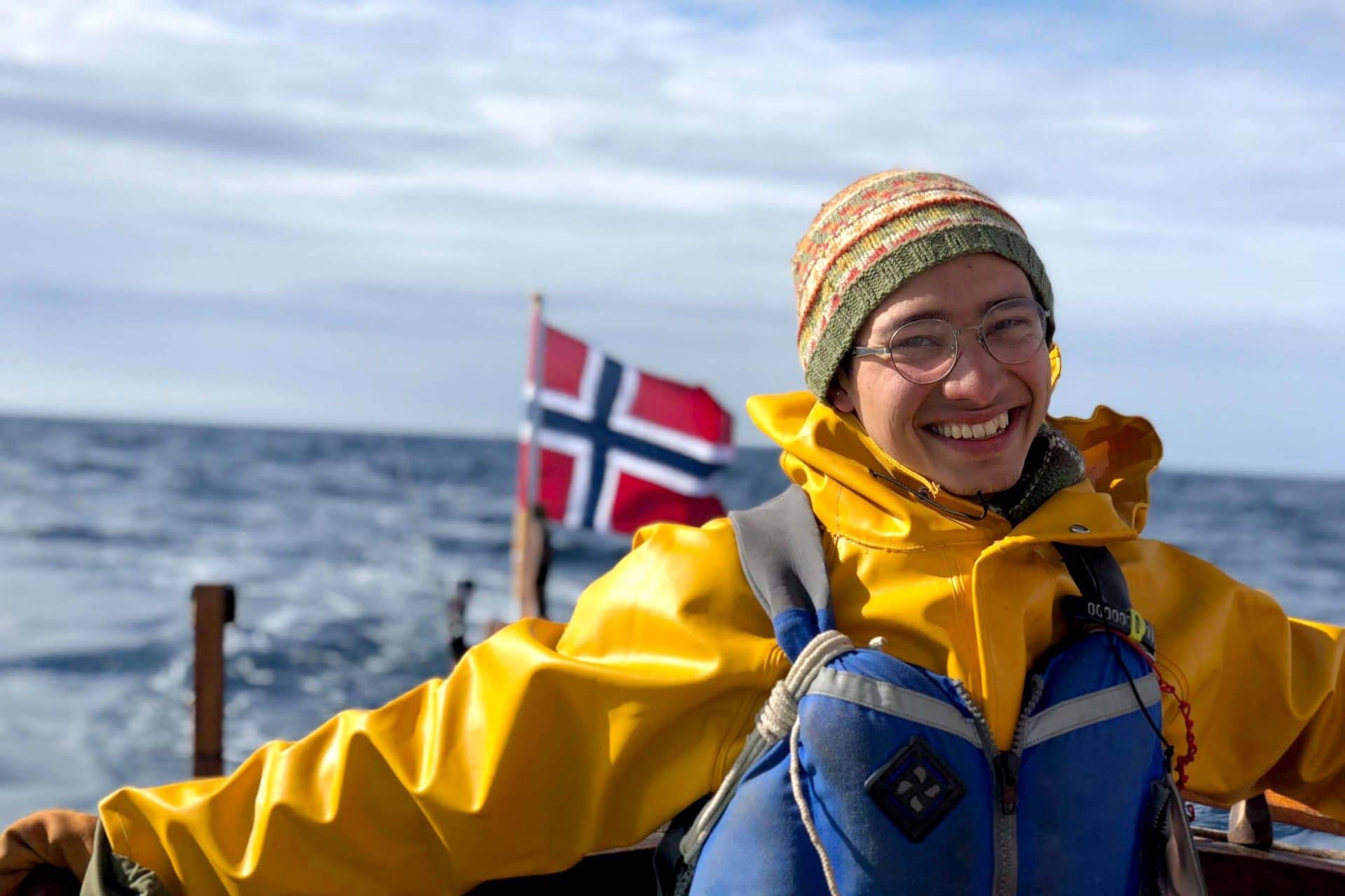 En glad elev med regntoy og flytevest styrer seilbaten i frisk vind. Det norske flagget i bakgrunnen.